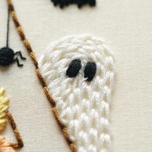 May include: A close-up of hand embroidery on off-white fabric featuring a white ghost with black thread eyes, a small black spider with thin legs, and a portion of a yellow and orange stitched element bordered by a brown thread line.