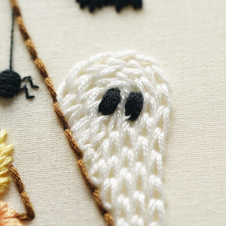 May include: A close-up of hand embroidery on off-white fabric featuring a white ghost with black thread eyes, a small black spider with thin legs, and a portion of a yellow and orange stitched element bordered by a brown thread line.