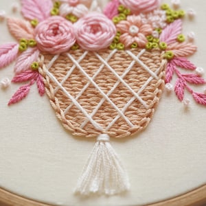 May include: Hand-stitched embroidery of a basket filled with pink roses, peach-colored flowers, and green accents. The basket is beige with a white lattice design and a white tassel. The embroidery is set against a cream-colored fabric.