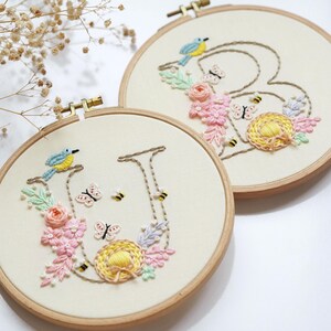 May include: Two embroidered hoops with floral and insect designs. One hoop features the letter "U" with a blue bird, butterflies, bees, and flowers. The other hoop has the letter "B" with similar elements. The embroidery is on a cream-coloured fabric.