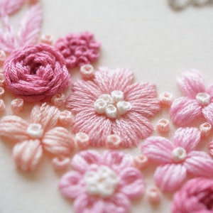 May include: Close-up of hand-stitched embroidery featuring pink and light pink flowers. The flowers include roses, daisies, and other floral designs. The embroidery is on a white fabric background.