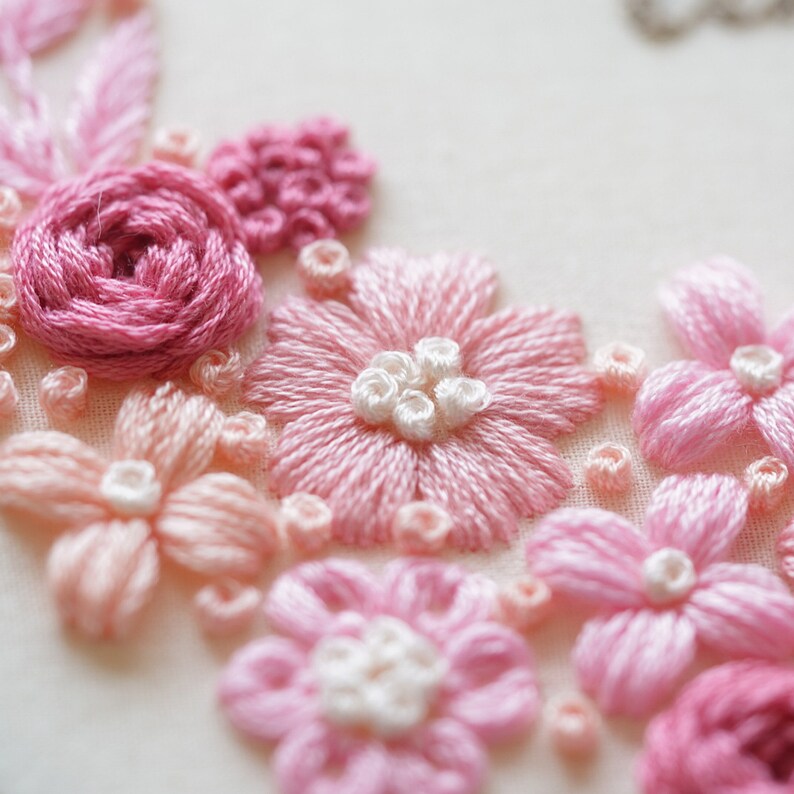 May include: Close-up of hand-stitched embroidery featuring pink and light pink flowers. The flowers include roses, daisies, and other floral designs. The embroidery is on a white fabric background.
