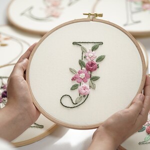 Op de afbeelding: Een houten borduurring houdt een cr&egrave;mekleurige stof vast met de geborduurde letter "J". De letter is versierd met roze en witte bloemenborduurwerk en groene bladeren. Andere ringen met geborduurde letters zijn zichtbaar op de achtergrond.