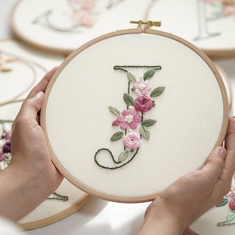 Op de afbeelding: Een houten borduurring houdt een cr&egrave;mekleurige stof vast met de geborduurde letter "J". De letter is versierd met roze en witte bloemenborduurwerk en groene bladeren. Andere ringen met geborduurde letters zijn zichtbaar op de achtergrond.