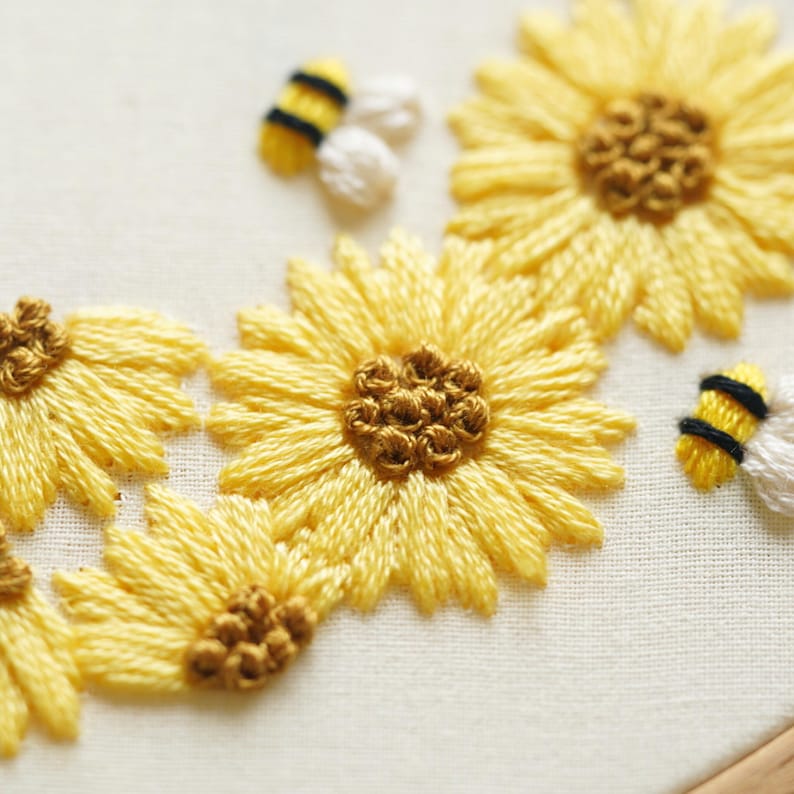 May include: Embroidered sunflowers and bees in a close-up shot. The sunflowers are yellow with brown centres, and the bees are yellow and black. The embroidery is on cream-coloured fabric, giving a handmade feel.