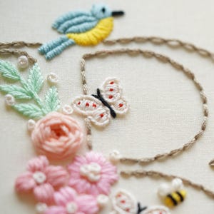 May include: Detailed embroidery showing a blue and yellow bird on a brown branch, a white butterfly with red spots, and pink flowers. The design also includes a small bee with yellow and black stripes, and green leaves.