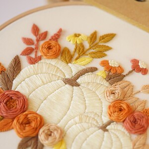 May include: An embroidery hoop featuring a hand-stitched design of two cream-colored pumpkins surrounded by a floral arrangement. The flowers and leaves are stitched in shades of orange, rust, brown, and yellow on a light cream fabric background.