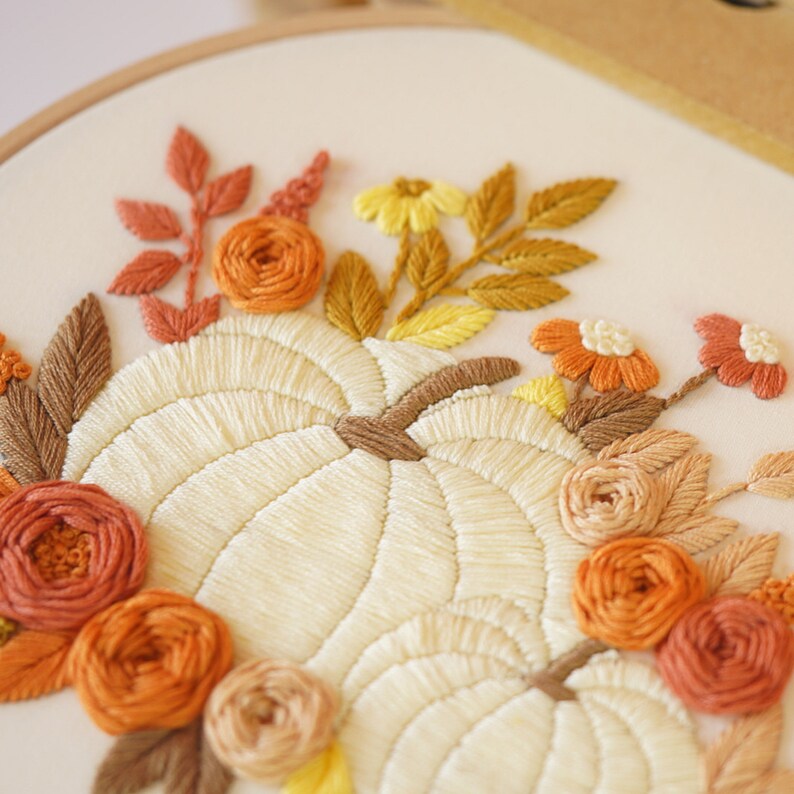 May include: An embroidery hoop featuring a hand-stitched design of two cream-colored pumpkins surrounded by a floral arrangement. The flowers and leaves are stitched in shades of orange, rust, brown, and yellow on a light cream fabric background.