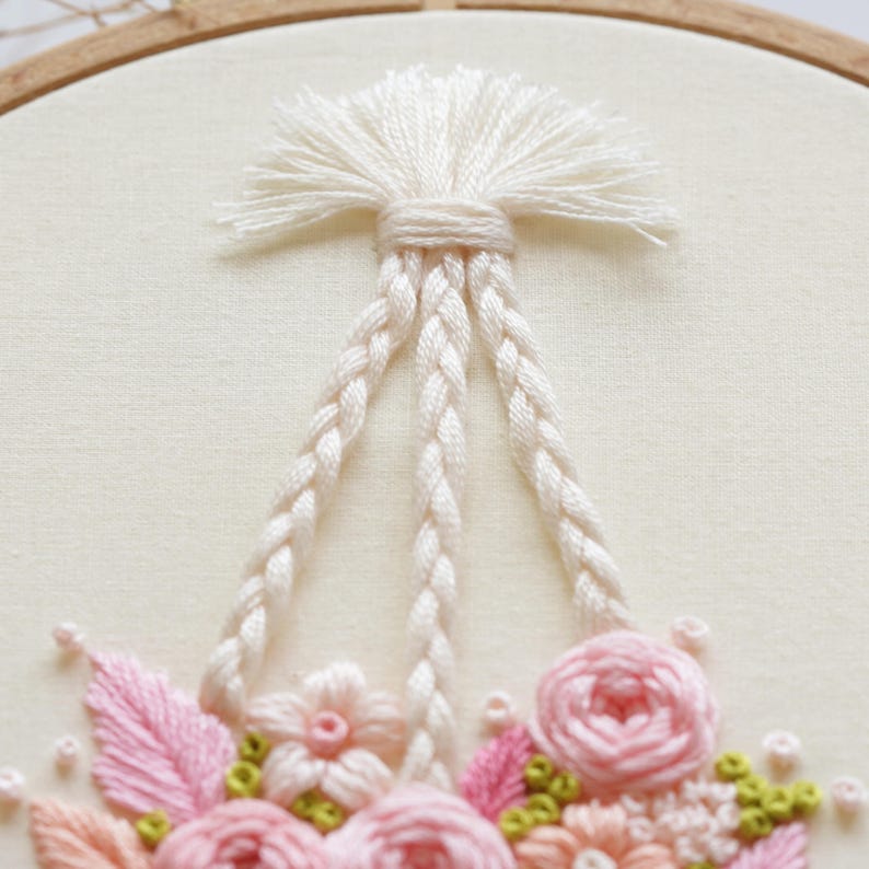 May include: Close-up of an embroidery hoop featuring a floral design. The design includes pink roses, leaves, and small flowers, with braided white cords and a tassel at the top. The embroidery is set against a cream-colored fabric.