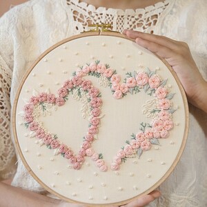 May include: A round embroidery hoop featuring a cream-colored fabric with a floral heart design. The hearts are formed with pink roses and green leaves. The hoop is held by a person.