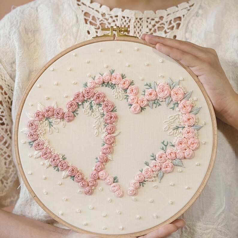 May include: A round embroidery hoop featuring a cream-colored fabric with a floral heart design. The hearts are formed with pink roses and green leaves. The hoop is held by a person.