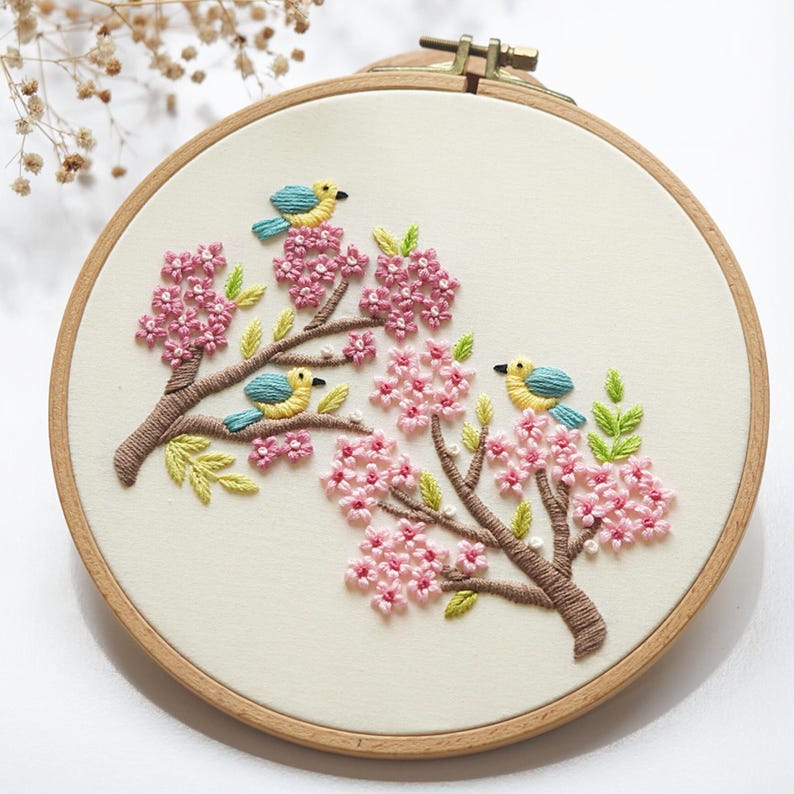 May include: Embroidered artwork featuring a wooden hoop with a cream-colored fabric. The design showcases pink cherry blossoms, brown branches, green leaves, and three blue and yellow birds. A charming piece of handmade decor.