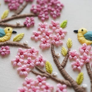 May include: Close-up of an embroidered design featuring pink cherry blossoms and yellow birds with blue wings. The embroidery is on a white fabric, with brown branches and green and yellow leaves. The design is detailed and colorful.