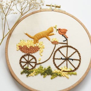 Pu&ograve; includere: Un telaio da ricamo con una scena cucita di un gatto arancione in un carretto pieno di fiori gialli, trainato da una bicicletta con telaio marrone. Un piccolo uccellino arancione &egrave; posato sul manubrio e fogliame verde &egrave; ricamato in basso.