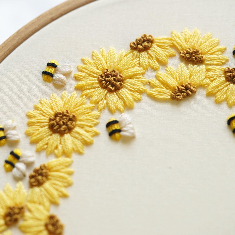 May include: Embroidered artwork showing yellow sunflowers and bees. The sunflowers have brown centres and yellow petals. The bees are white, yellow, and black. The embroidery is on cream-coloured fabric, set in a wooden hoop.