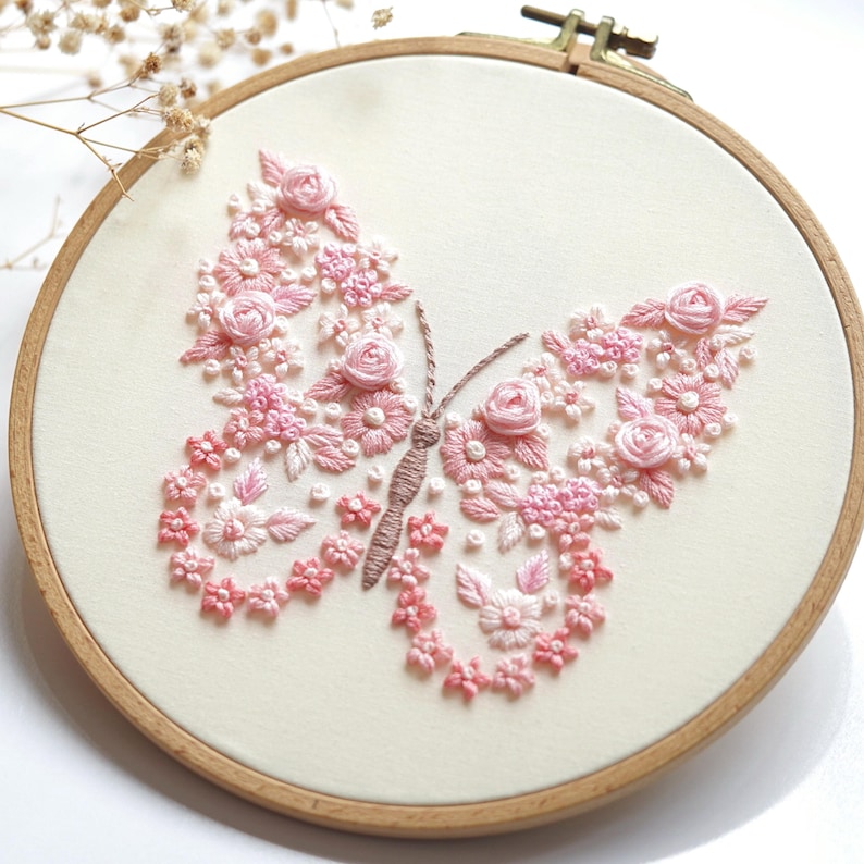 May include: An embroidered butterfly design on a cream-coloured fabric, set within a wooden hoop. The butterfly is formed from pink and light pink flowers and leaves. The craftwork is a delicate piece of textile art.
