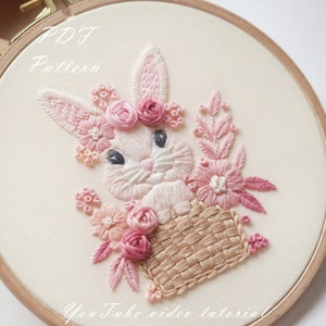 May include: Embroidered hoop art featuring a white bunny with pink ears and a pink flower crown, sitting in a woven basket with pink flowers. The text "PDF Pattern" and "YouTube video tutorial" are also included.