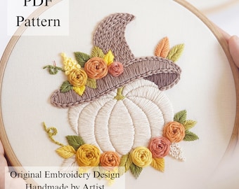 May include: An embroidery hoop containing a white fabric panel with a stitched design of a white pumpkin wearing a brown witch hat. The pumpkin is decorated with yellow, orange, and green floral accents. Text reads PDF Pattern and Original Embroidery Design.