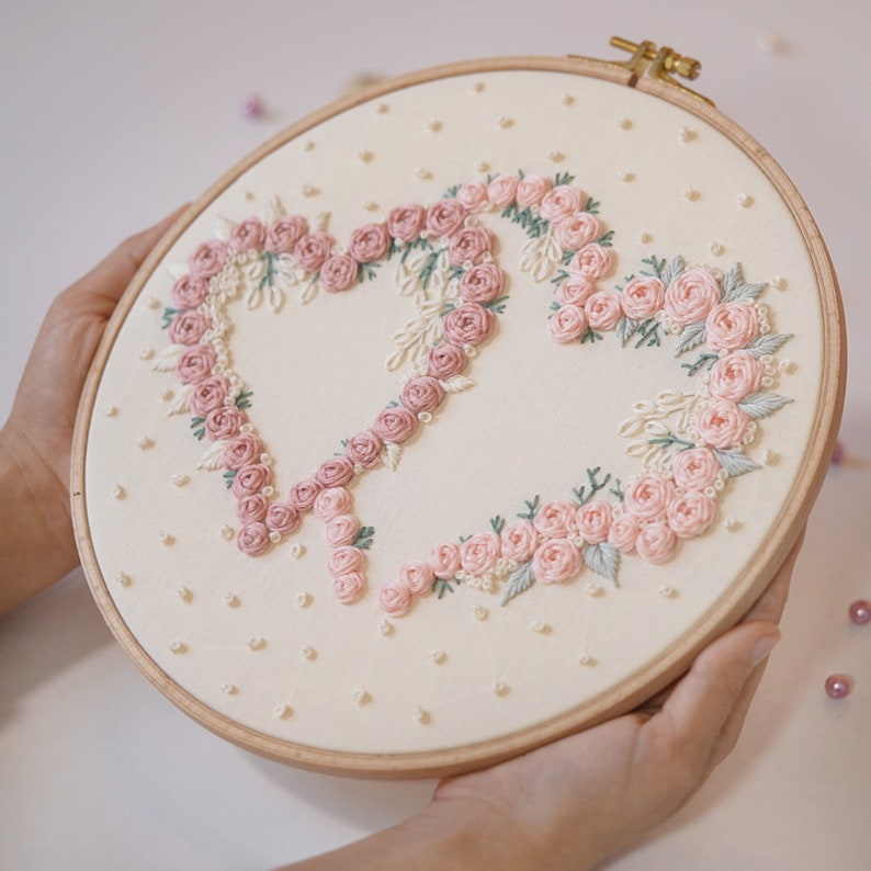 May include: An embroidered hoop art piece featuring two hearts made of pink roses and green leaves on a cream-colored fabric. The embroidery is set in a wooden hoop, with small white beads scattered across the background.