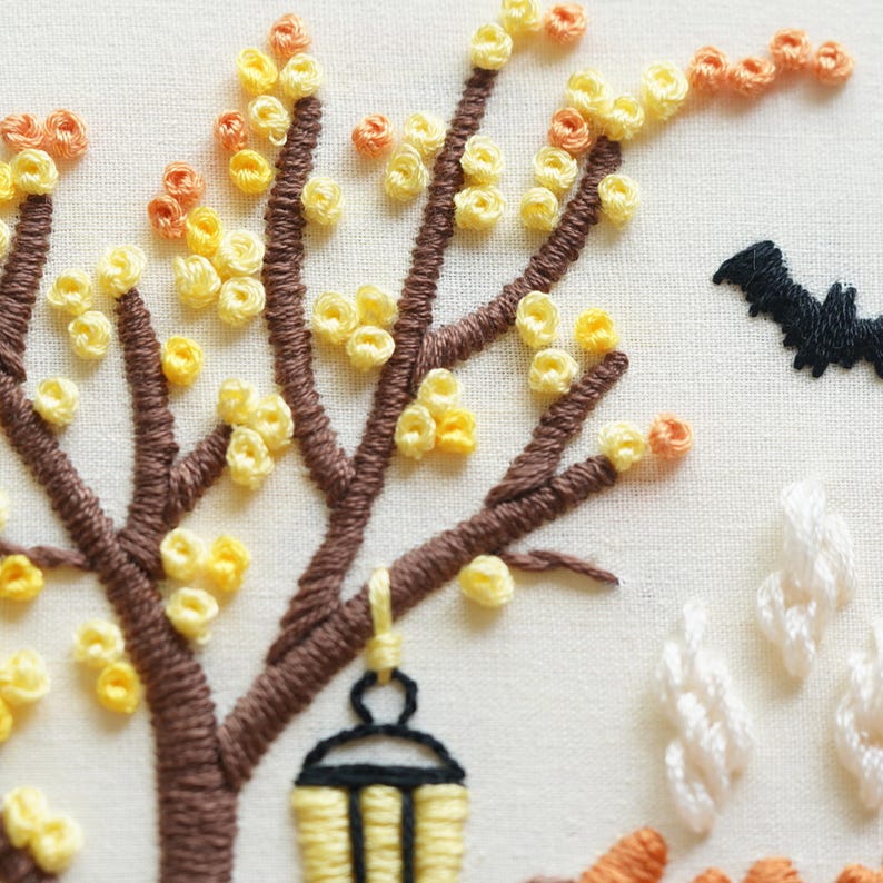 May include: An embroidery piece on off-white fabric featuring a brown tree with yellow and orange French knot leaves. A black lantern hangs from a branch. To the right, a black bat is stitched, along with white textured clouds or smoke shapes.