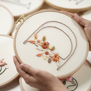 Op de afbeelding: Een close-up van een bloemenborduurring die in handen wordt gehouden. De borduursels tonen de letter "A" met een bloemenontwerp in tinten oranje, wit en geel. De ring is van hout en de stof is cr&egrave;mekleurig. Andere borduurringen zijn zichtbaar op de achtergrond.