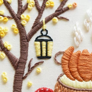 May include: An embroidery piece on off-white fabric featuring a brown tree with yellow French knot leaves. A yellow and black lantern hangs from a branch. Below, an orange pumpkin sits near a red mushroom, with white stitched steam rising upward.