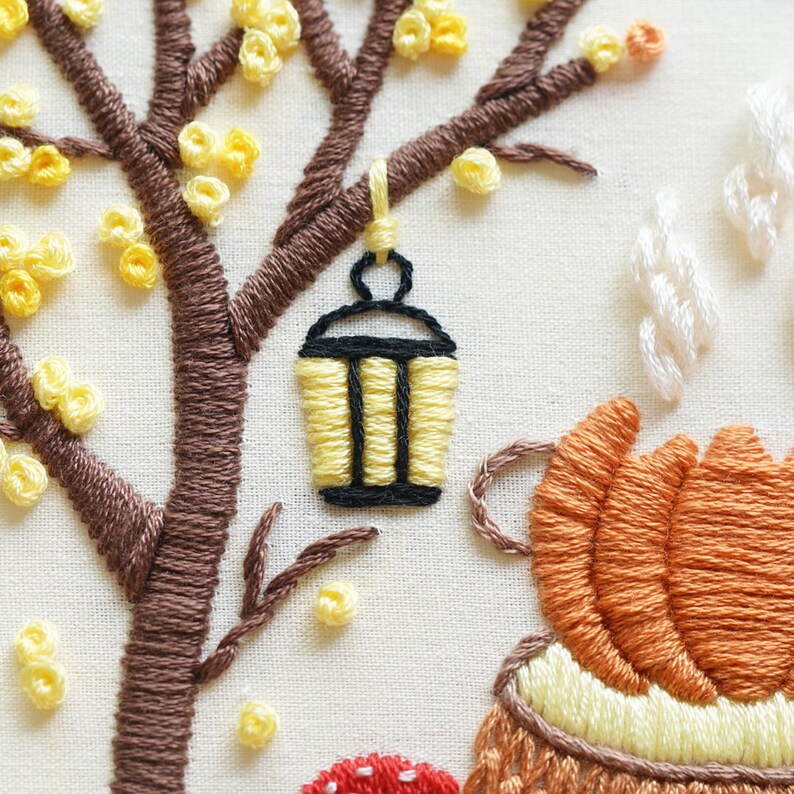 May include: An embroidery piece on off-white fabric featuring a brown tree with yellow French knot leaves. A yellow and black lantern hangs from a branch. Below, an orange pumpkin sits near a red mushroom, with white stitched steam rising upward.