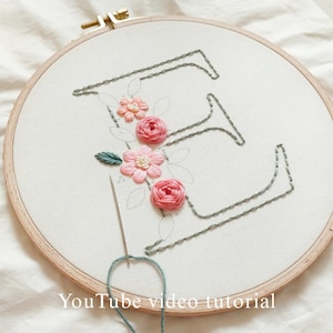 Op de afbeelding: Een borduurring met de letter "E" gestikt in grijze draad, versierd met roze en perzik bloemen en groene bladeren. Een naald met blauwe draad is in de stof gestoken. De tekst "YouTube video tutorial" staat onderaan.