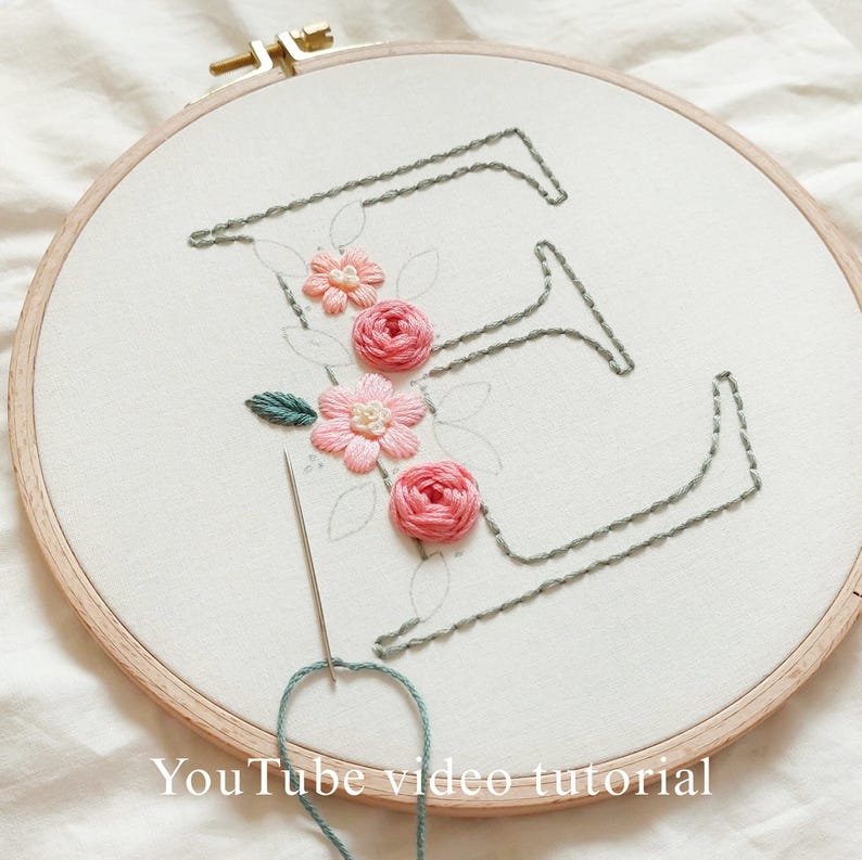 Op de afbeelding: Een borduurring met de letter "E" gestikt in grijze draad, versierd met roze en perzik bloemen en groene bladeren. Een naald met blauwe draad is in de stof gestoken. De tekst "YouTube video tutorial" staat onderaan.