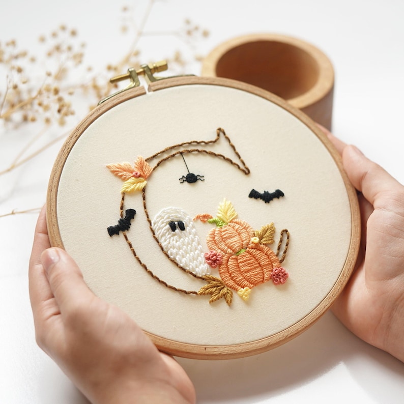 May include: A circular wooden embroidery hoop containing off-white fabric with a hand-stitched letter C. The design features a white ghost, orange pumpkins, yellow and brown autumn leaves, two black bats, and a small black spider hanging from the letter.
