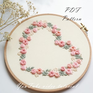 May include: A heart-shaped embroidery design featuring pink roses and green leaves, set within a wooden embroidery hoop. The text "PDF Pattern" and "YouTube video tutorial" are visible. The design is on a cream-colored fabric.