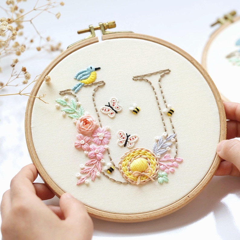 May include: Embroidered hoop art with a floral design and the letter "U". The artwork includes a blue bird, pink and peach flowers, butterflies, and bees. The embroidery is set in a wooden hoop.