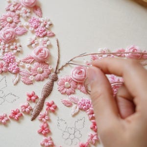 May include: A hand is shown embroidering a pink floral butterfly design on cream fabric. The embroidery features various pink flowers and leaves, with a detailed butterfly body. A needle and thread are in use, showcasing the craft process.