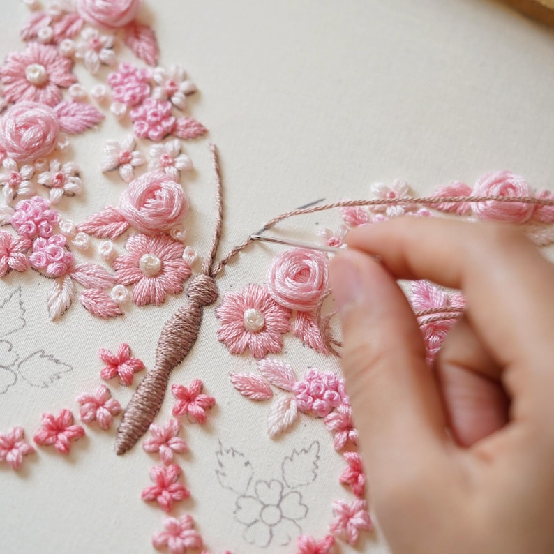 May include: A hand is shown embroidering a pink floral butterfly design on cream fabric. The embroidery features various pink flowers and leaves, with a detailed butterfly body. A needle and thread are in use, showcasing the craft process.