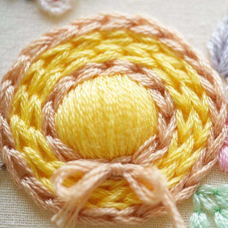 May include: Close-up of a hand-stitched embroidery piece. The design features a central yellow circle surrounded by concentric rings of yellow and peach-coloured thread. A small peach-coloured bow is at the bottom.