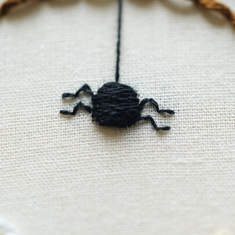 May include: A black spider embroidered with thick thread on a cream-colored fabric background. The spider is depicted with a round body and multiple legs, suspended by a single vertical black thread extending from the top of its body.