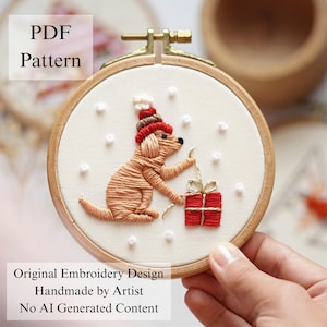 May include: A round wooden embroidery hoop containing a cream-colored fabric with a hand-stitched design of a tan dog wearing a red and brown winter hat, reaching toward a red gift box with gold ribbon, surrounded by small white French knots.