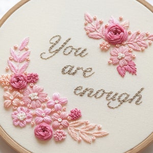 May include: Hand-stitched embroidery hoop featuring the phrase "You are enough" in cursive script. The design includes pink and peach-colored floral arrangements. The embroidery is set against a cream-colored fabric background.