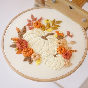 May include: An embroidery hoop containing a white fabric panel with a hand-stitched design of two cream-colored pumpkins surrounded by a floral arrangement of orange, rust, yellow, and brown thread-stitched flowers and autumn-toned leaves.