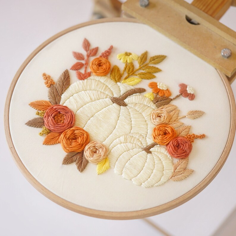 May include: An embroidery hoop containing a white fabric panel with a hand-stitched design of two cream-colored pumpkins surrounded by a floral arrangement of orange, rust, yellow, and brown thread-stitched flowers and autumn-toned leaves.