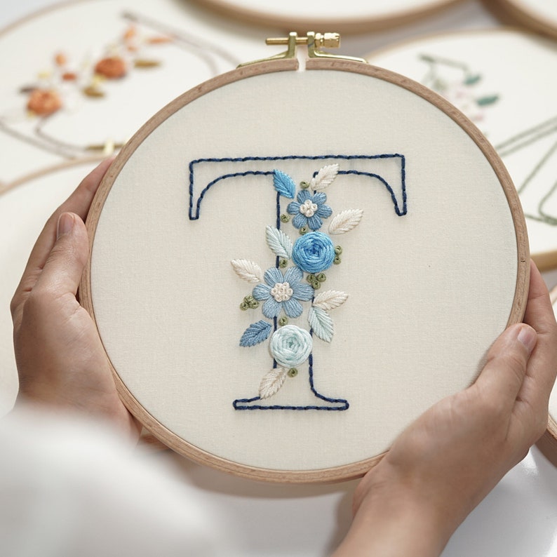 Op de afbeelding: Een handgeborduurde borduurring met de letter 'T' in marineblauwe draad, versierd met licht- en middelblauwe bloemen- en blad details. De borduursels zijn geplaatst op een cr&egrave;mekleurige stof, omlijst door een houten ring.