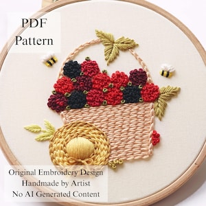Könnte beinhalten: Ein Stickrahmen mit einem Stoffstück, das ein gesticktes Motiv eines Korbes voller roter und schwarzer Beeren zeigt. Das Design umfasst einen gelben Strohhut, grüne Blätter und zwei kleine Bienen. Text: PDF Pattern, Original Embroidery Design.