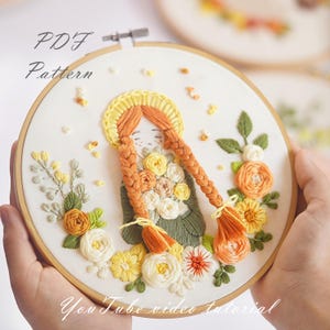 Op de afbeelding: Borduurwerk in een borduurring met een meisje met lange oranje vlechten en een bloemenkrans. Het meisje draagt een groene jurk en houdt een boeket bloemen vast. De ring is omgeven door een witte achtergrond met de tekst "PDF Pattern" en "YouTube video tutorial".