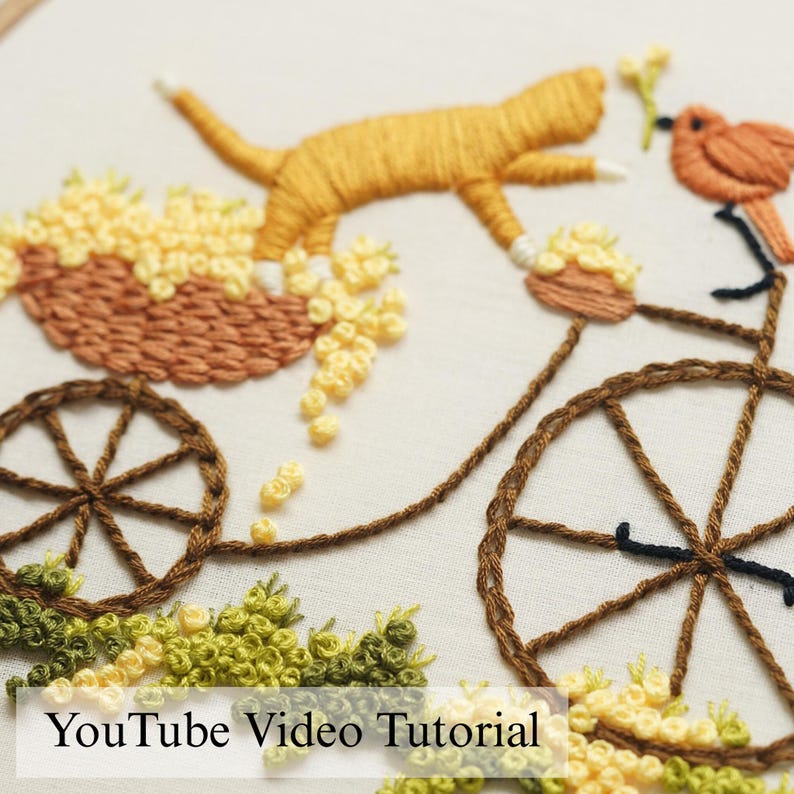 Pu&ograve; includere: Un ricamo su tessuto bianco raffigurante un gatto dorato che salta sopra un carretto marrone pieno di fiori gialli. &Egrave; visibile una bicicletta marrone con un piccolo uccellino arancione sul manubrio, con fogliame verde e fiori gialli. YouTube Video Tutorial.