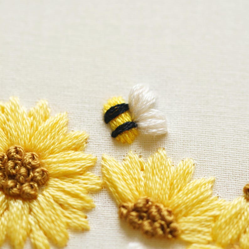 May include: Close-up of embroidered sunflowers and a bee. The sunflowers are yellow with brown centres, and the bee has a yellow body with black stripes and white wings. The embroidery is on a cream-coloured fabric.