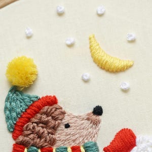 K&ouml;nnte beinhalten: Eine Stickerei auf cremefarbenem Stoff zeigt einen Igel mit einer t&uuml;rkisfarbenen Strickm&uuml;tze mit gelbem Bommel, rotem Rand und einem rot-t&uuml;rkisen Schal. Dar&uuml;ber sind ein gelber Halbmond und kleine wei&szlig;e Schneeflocken aufgestickt.