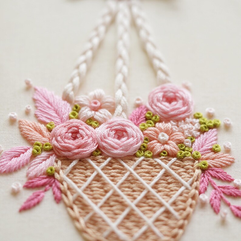 May include: Embroidered basket filled with pink and peach flowers, green accents, and pink leaves. The basket is tan with a white crisscross pattern. The basket has white braided handles. The background is a light cream color.