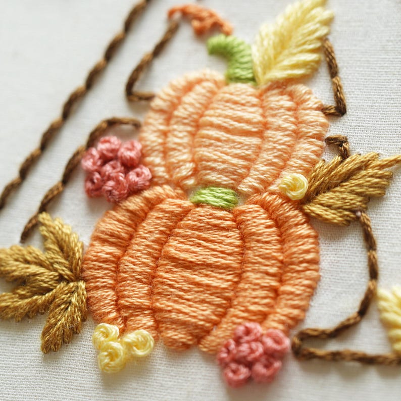 May include: An embroidery design on white fabric featuring two stacked orange pumpkins with light green stems. The pumpkins are surrounded by golden-yellow leaves, small clusters of pink French knots, and brown stitched vines framing the composition.