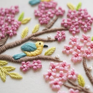May include: Embroidered artwork featuring a yellow and blue bird perched on a brown branch. Pink cherry blossoms and green leaves surround the bird. The embroidery is detailed and colorful, with a focus on texture and depth.