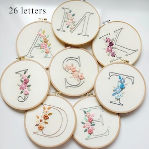 Op de afbeelding: Een verzameling geborduurde letters, elk in een houten hoepel. De letters zijn omrand met zwarte draad en versierd met kleurrijke bloemenborduurwerken. De tekst "26 letters" is zichtbaar bovenaan.